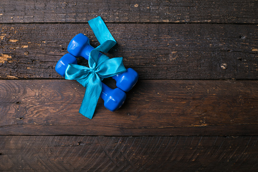 two blue dumbbell with a gift bow and blank paper tag, heart on wooden planks, sport holiday concept