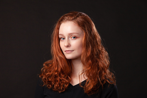 Close up studio portrait of 18 year old woman with long curly red hair on black background