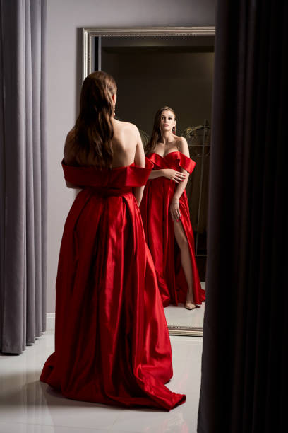 young beautiful girl wearing an off-the-shoulder full-length crimson red satin slit prom ball gown. model looking in mirror. fitting room in dress hire service. - women cheerful vertical 20s imagens e fotografias de stock