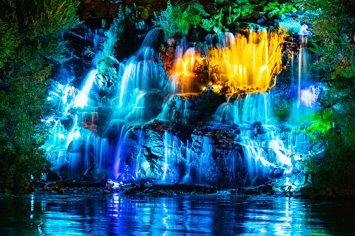 Waterfall in neon color. Flowing water illuminated by multicolored light. Night landscape with a colorful waterfall near the river