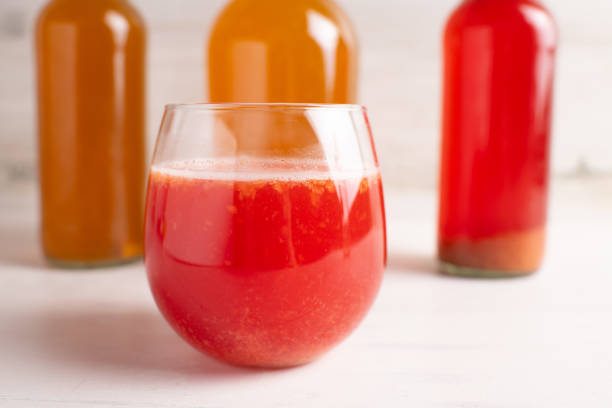 Strawberry Kombucha in small glass with pineapple and other bottled flavors in the background.  Selective Focus is in the glass.