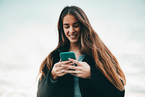 Young cheerful smiling happy woman checking messages on her smart phone outdoors in wintertime, Real People Outdoor Millenial Youth Lifestyle Portrait.