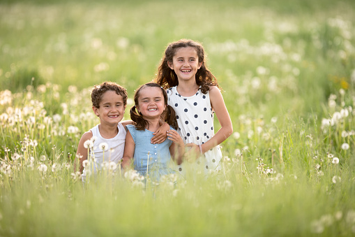 A trio of siblings all happy and smiling together as they are standing in a field full of dandelions on a bright summer day.