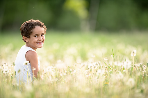 An adorable elementry age boy smiles widely at the camera as he sits in a field full of dandelions on a bright summer day.