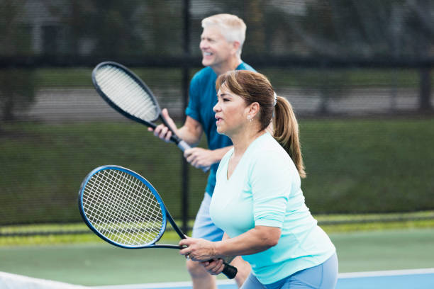 Couple playing tennis, focus on Hispanic woman A couple playing tennis together. They are a doubles team. The focus is on the Hispanic woman standing at the net in the foreground, waiting for a volley. The is a mature woman in her 50s. Her partner is a senior man in his 60s. tennis senior adult adult mature adult stock pictures, royalty-free photos & images