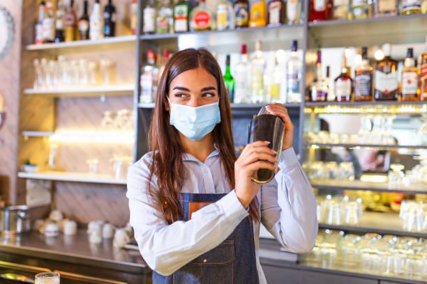 istock Young female worker at bartender desk in restaurant bar preparing coctail with shaker. Beautiful young woman behind bar wearing protective face mask 1294235012