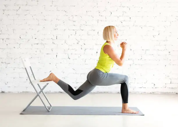 Workout with props. Fit adult caucasian woman practice lunges with one leg on a chair on a mat in loft white studio indoor, selective focus. Yoga, fitness, trainer, sport, healthy lifestyle, concept.