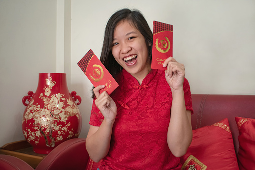 Portrait shot of happy young Chinese Indonesian woman with modern red cheongsam showing her red envelope angpao, sitting on the living room