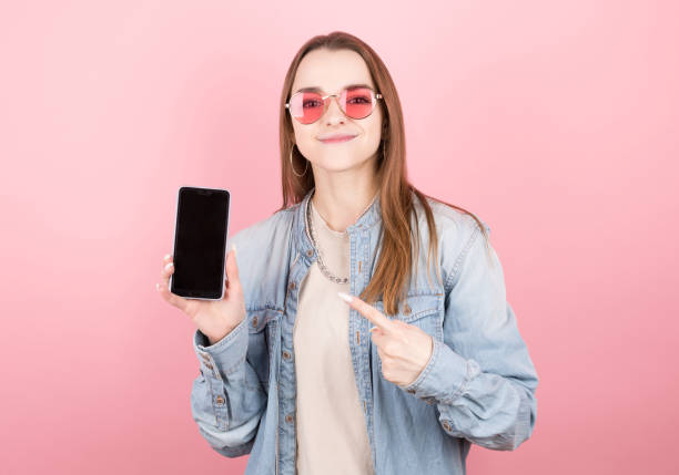 istock Surprised happy brunette in denim jacket showing blank smartphone screen and pointing at it while looking at camera with smile on pink background 1294284577