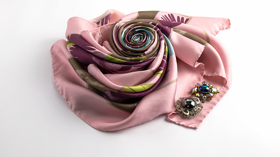 Old brooches with sparkling crystals lie on the floral pink and purple  silk scarf folded in the shape of a rose. Isolated object on the white background.