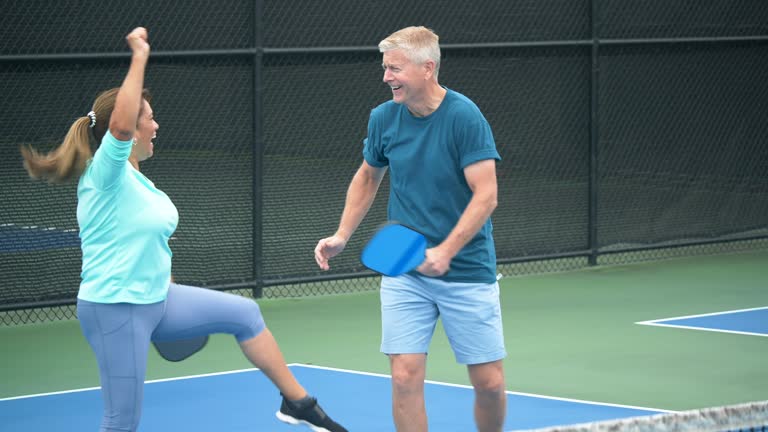 A couple playing pickleball together. They are a doubles team. The Hispanic woman, in her 50s, hits a winning shot with her pickleball paddle. Her partner, a senior man in his 60s, gives her a high-five as they celebrate.