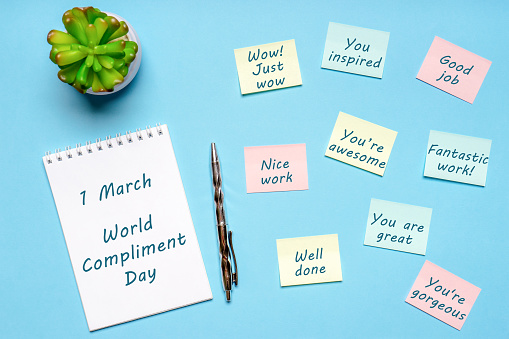 Happy World Compliment Day. Office desk with plant, notebook, pen and paper slips with compliments text for office worker such as GOOD JOB. Greeting card for world compliment day. Flat lay, top view.