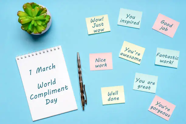 Photo of Happy World Compliment Day. Office desk with plant, notebook, pen and paper slips with compliments text for office worker such as GOOD JOB. Greeting card for world compliment day. Flat lay, top view