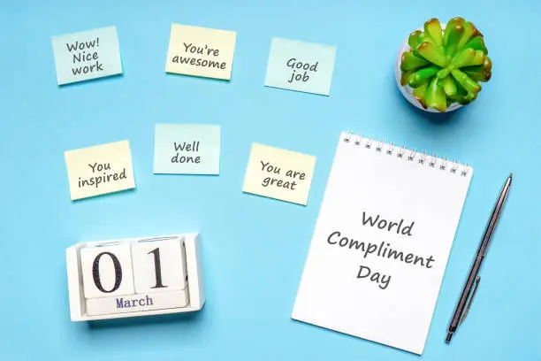 Photo of Happy World Compliment Day. Office desk with plant, notebook, pen and paper slips with compliments text for office worker such as GOOD JOB. Calendar date 1 March, greeting card. Flat lay, top view