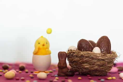 Bird nest with Chocolate eggs and decorative chick on table full of easter almonds against white background
