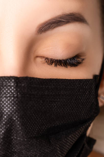 istock Eyelash Extension Procedure. Beautiful Woman with Extreme Long False Eyelashes. Makeup, Cosmetics. Beauty, Skincare. Woman in medical mask 1294303568