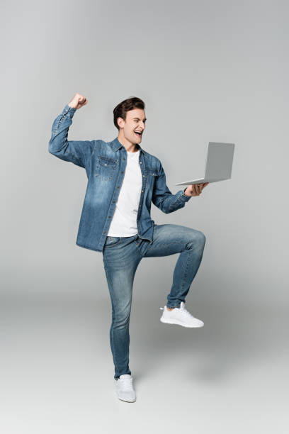 istock Cheerful man showing yeah gesture while holding laptop on grey background 1294313811