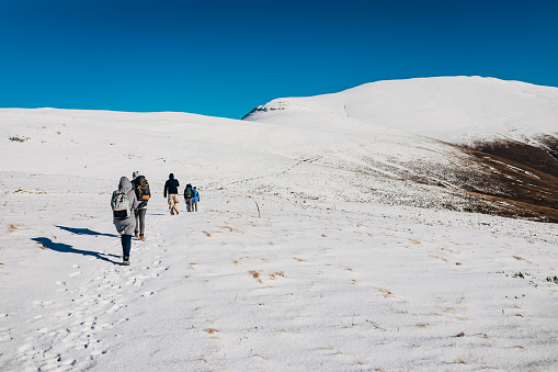 Group of male and female hikers wearing warm clothes and backpacks, hiking and reaching the top of the snowy mountain during sunny winter day