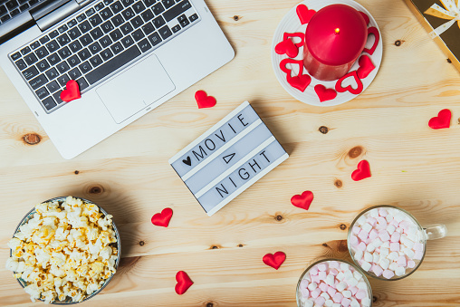 St. Valentine's day Movie night concept. Movie night message on board, laptop, popcorn, red candle, gift, hearts decor, two cups of cocoa with marshmallows for a couple. Cozy holiday plans for lovers
