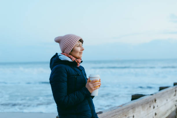 暖かいジャケットと帽子をかぶった笑顔の女性で、再利用可能なカップを持ち、熱い飲み物が桟橋に立ち、冬の海の景色を楽しんでいます。シンプルな喜びとローカル旅行、デジタルデトッ� - winter women zen like photography ストックフォトと画像