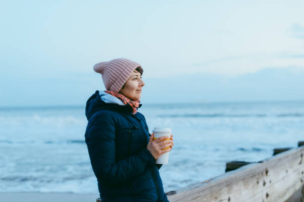 smiling woman in a warm jacket and hat with reusable cup with hot drink standing on the pier and enjoying the winter sea view. simple pleasures and local traveling, digital detox concepts. copy space. - winter women zen like photography imagens e fotografias de stock