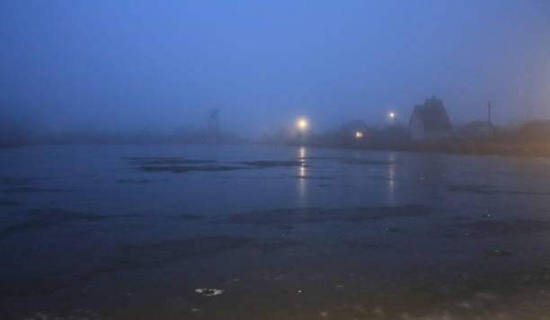 vista de uma casa solitária na neblina no início da manhã. o lago está coberto de gelo na neblina. lâmpada solitária e janela na neblina - street light fog morning country road - fotografias e filmes do acervo