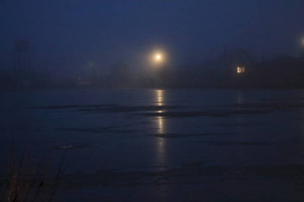 vista de uma casa solitária na neblina no início da manhã. o lago está coberto de gelo na neblina. lâmpada solitária e janela na neblina - street light fog morning country road - fotografias e filmes do acervo