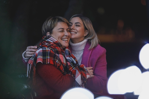 Candid shot of happy young woman and her mature mother having a bonding moment while enjoying magical winter evening at the park, cuddling and enjoying spending time together.