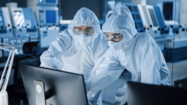 Research Factory Cleanroom: Engineer and Scientist Wearing Coveralls Talk and Work on Computer, Use Microscope to Inspect Motherboard Microprocessor, Develop Electronics for Medical Electronics Research Factory Cleanroom: Engineer and Scientist Wearing Coveralls Talk and Work on Computer, Use Microscope to Inspect Motherboard Microprocessor, Develop Electronics for Medical Electronics cleanroom photos stock pictures, royalty-free photos & images