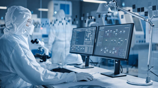 istock Electronics Factory Cleanroom: Engineer / Scientist in Coveralls Works on Computer, Screen Shows Infographics and Software System Control UI, Developing Electronics for Medical Electronics 1294339638