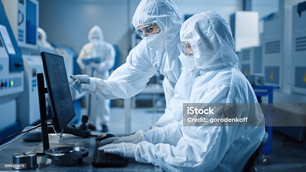Factory Cleanroom: Engineer and Scientist Wearing Coveralls and Masks Have Discussion, Use Computer Showing Infrastructure System Control. In Background CNC Machinery, Electronics Equipment Pharmaceutical Industry Stock Photo