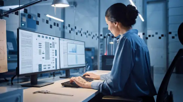 Photo of Factory Office: Portrait of Beautiful and Confident Female Industrial Engineer Working on Computer, on Screen Industrial Electronics Design Software. High Tech Facility with CNC Machinery