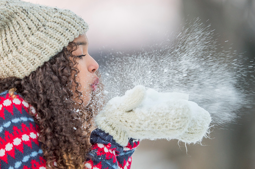 A mixed race female teenager blows snow away from her hands as she enjoys nature in the cold winter time.