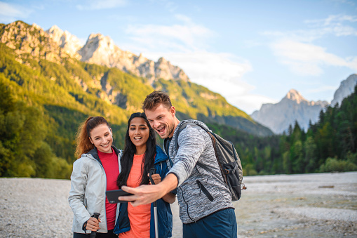 Low angle view of three young male and female friends pausing from alpine hike to smile for selfie near Kranjska Gora with Julian Alps in background.