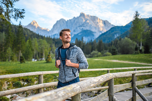 Close-up of male hiker in early 30s enjoying view while crossing pedestrian bridge near Lake Jasna with afternoon light shining on Mt. Prisojnik in background.
