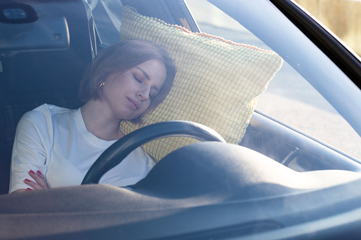 Tired young woman driver asleep on pillow inside her car, resting after long hours driving. Fatigue. Sleep deprivation.