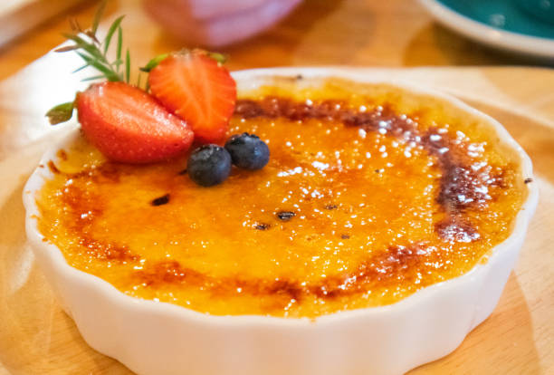 a golden yellow creme brulee topped with crunchy brown sugar topped with strawberries and blueberries to be included in a white glass plate on the dining table after eating. - custard creme brulee french cuisine crema catalana imagens e fotografias de stock