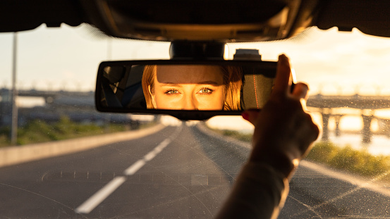 Cheerful joyful woman adjusting mirror while sitting in her car, looking in reflection at camera. Sunset time.
Emotions from driving car