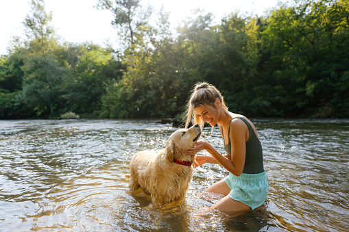 Photo of a smiling young woman and her dog bathing in the river; having a great time and enjoying the sunny afternoon outdoors.