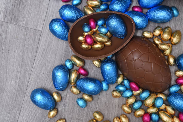istock Pile or group of multi colored and different sizes of colourful foil wrapped chocolate easter eggs in pink, blue, gold and green. Large halves of a brown milk chocolate egg have mini eggs inside, on a grey wooden background. 1294400133