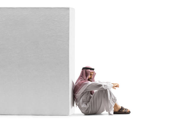 Sad saudi arab man sitting on the floor and leaning on a wall isolated on white background