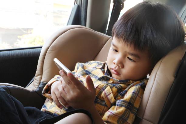 Asian boy looking at smartphone screen. Child sitting on car seat and watch video cartoon on mobile phone. Kid holding cellphone in his hand. stock photo
