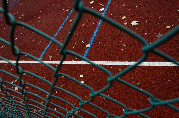 nahaufnahme des metallzauns gegen dunkelroten sportplatz und weiße, blaue linien darauf - athleticism multi colored red brown stock-fotos und bilder