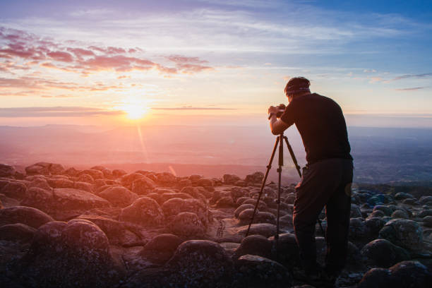mann fotograf hält eine kamera, um die natur sonnenaufgang auf berglandschaft zu schießen. touristen fotografieren die natur des sonnenuntergangs mit der kamera auf einem stativ mit kopierraum. - fotograf stock-fotos und bilder
