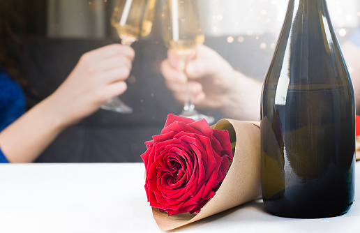 couple clink glasses with champagne in the background, a bottle of champagne and a rose in the foreground