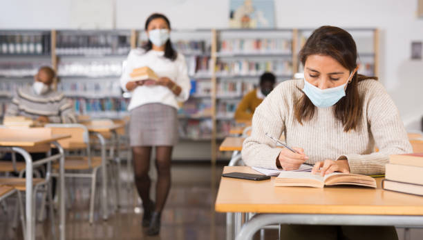 istock Young hispanic woman in protective mask reading books in library 1294410111