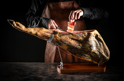 Iberian ham serrano ham cutting slice male hands and knife on dark moody background low key black hoof