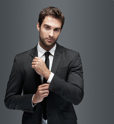 Confident young man doing his cufflinks up and looking at the camera, isolated on grey - copyspace