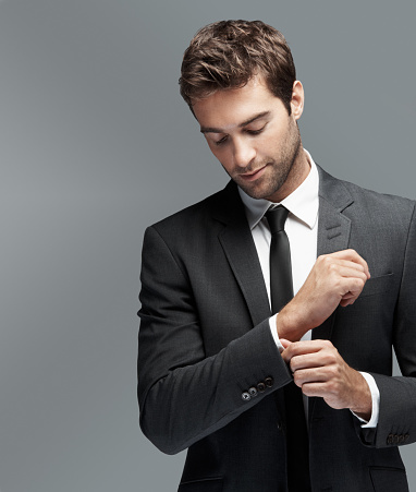Serious young man looking down as he is doing his cufflink, isolated on grey - copyspace