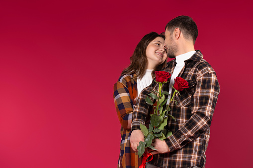 A loving couple with flowers on a red background look cute into each other's eyes. Empty side space. High quality photo